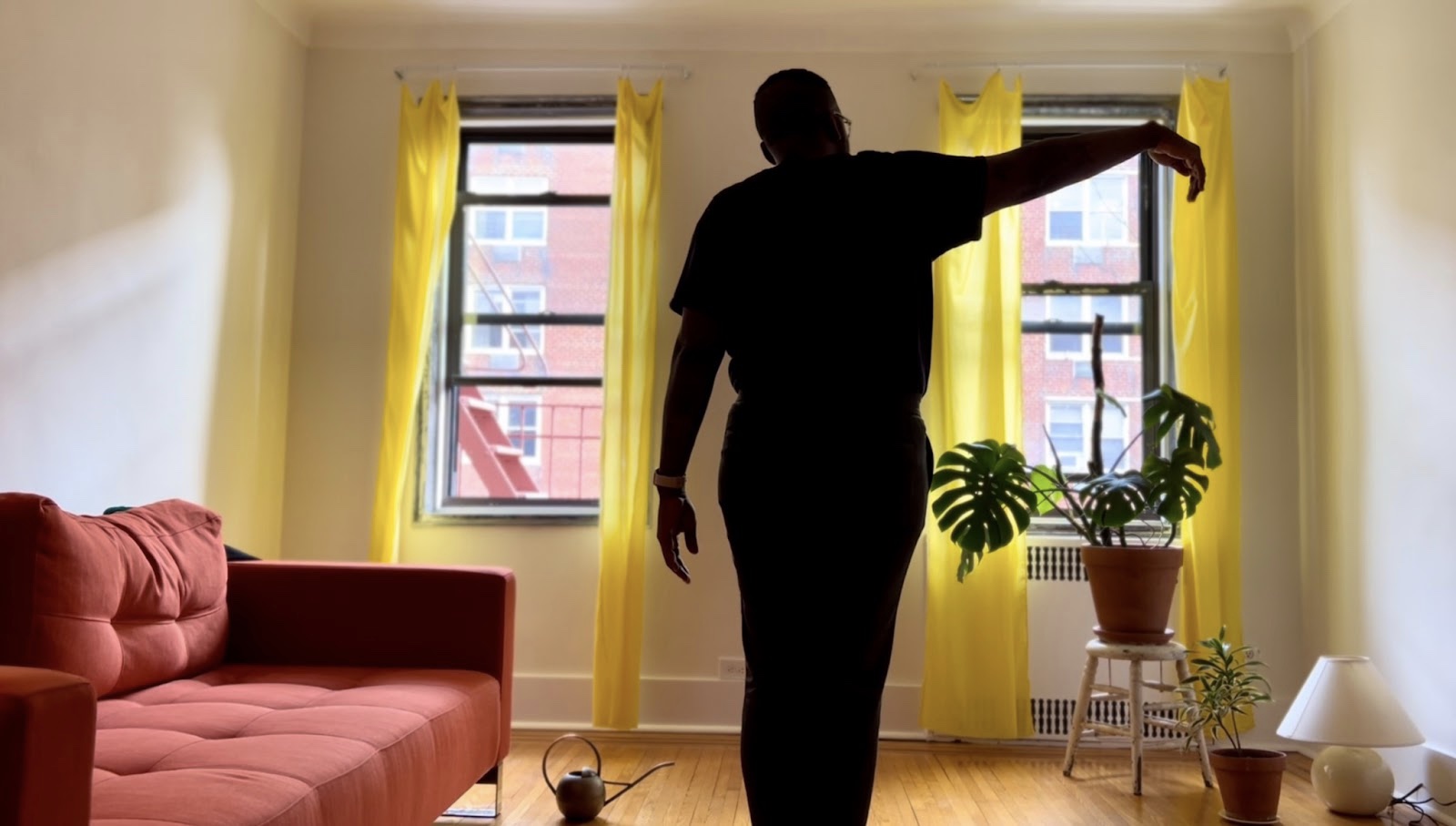 Inside the same apartment, we are now positioned further back in the room. On the left there is a red sofa, a watering can sits beside it on the floor. In the centre of the image we can see the silhouette of JT Green, they are facing the window with the right arm raised in an elegant line. On the floor to their right there is a lamp on the floor, another plant and a full view of the Cheese Plant perched on top of a stool.