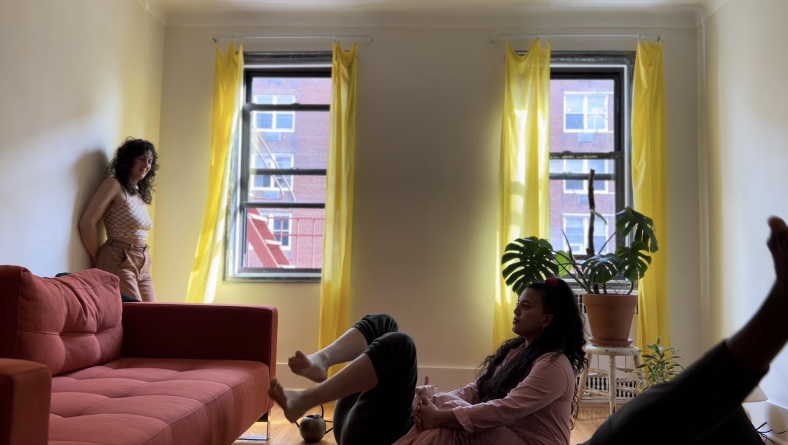 The third image holds the same view of the room but this time we can see four people in the image - someone stands by the window on the left hand side, their back pressed up against the wall, watching the scene. In the centre two people sit on the floor - whilst only the legs and stomach are visible for one as they lie on their back, another sits in front, calmly facing the sofa, their hands gently folded around their knees. They form a kind of optical illusion as if they are part of a single body. In the foreground we can infer that someone is lying on the floor holding their leg aloft, as the calf and toes are just about visible.
